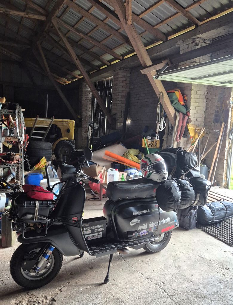 Lambretta grigia carica di bagagli parcheggiata in un garage rustico, tra attrezzi e materiali da lavoro.