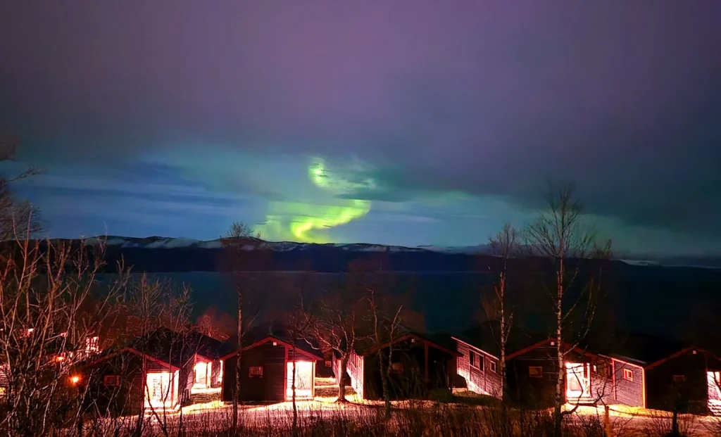 Aurora boreale verde sopra un lago circondato da montagne in Svezia, con chalet illuminati in primo piano.