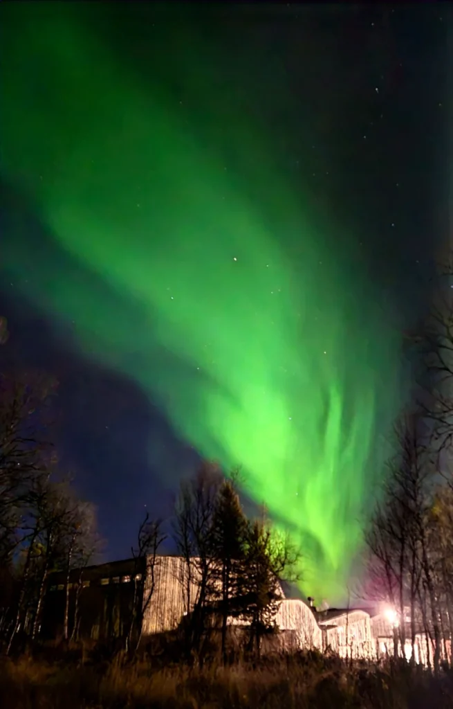Intensa aurora boreale verde illumina il cielo stellato sopra gli edifici e gli alberi della Lapponia svedese.