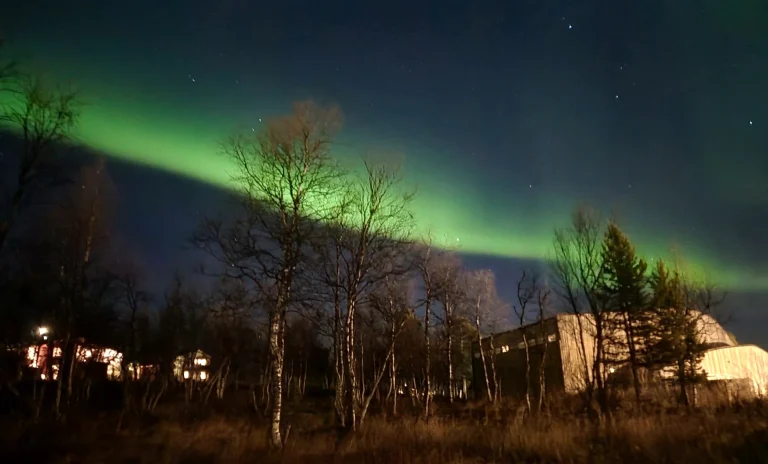Aurora boreale verde nel cielo notturno della Svezia, sopra gli alberi e le strutture illuminate ai piedi delle montagne.