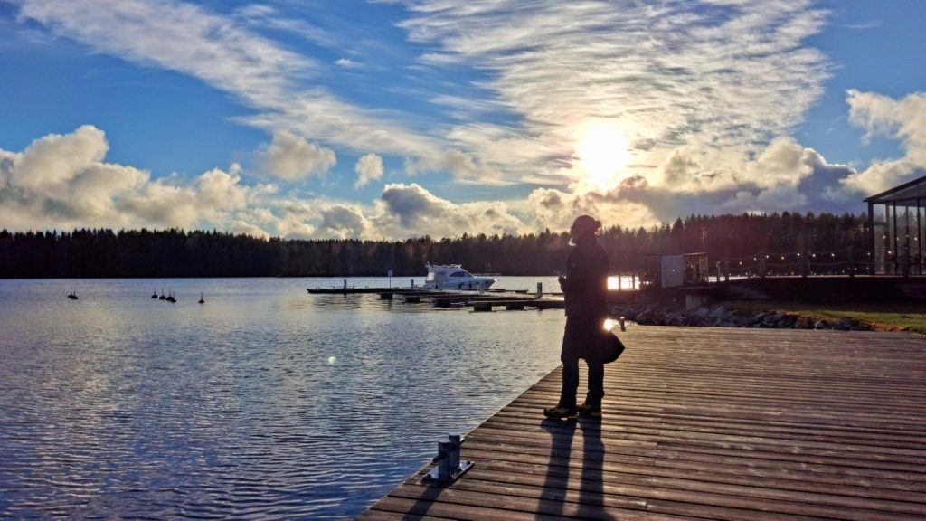 Persona su un pontile di legno al tramonto, con lago, imbarcazioni e foreste sullo sfondo nella regione dei laghi finlandesi.