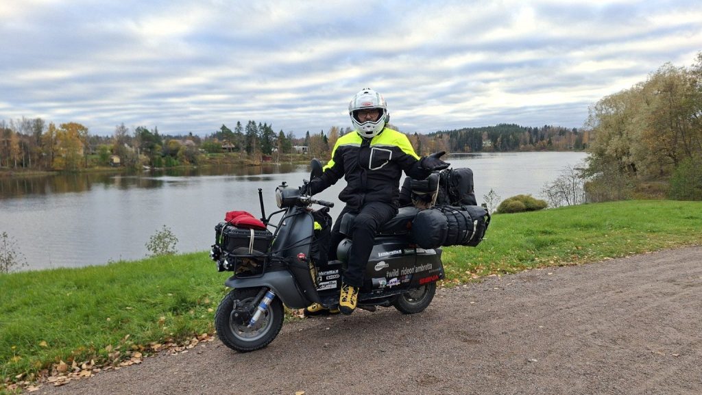 Filippo sulla Lambretta Xena carica di bagagli davanti a un lago a Kouvola, in Finlandia, durante The Great North.