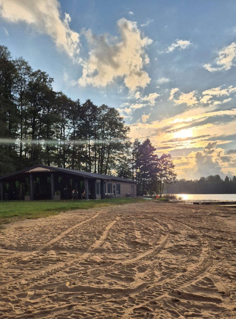 Spiaggia sabbiosa sul lago Szelment Wielki, con edificio sulla riva, alberi e sole al tramonto.