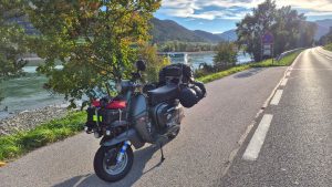 Lambretta grigia carica di bagagli lungo una strada che costeggia il Danubio in Austria, con un battello turistico sul fiume.