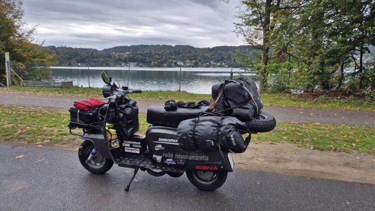 Lambretta grigia carica di bagagli da viaggio parcheggiata davanti a un lago in Austria, con colline boscose sullo sfondo.