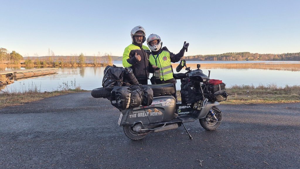 Francesca e Filippo con la Lambretta Xena carica di bagagli davanti a un lago circondato da boschi autunnali in Finlandia.