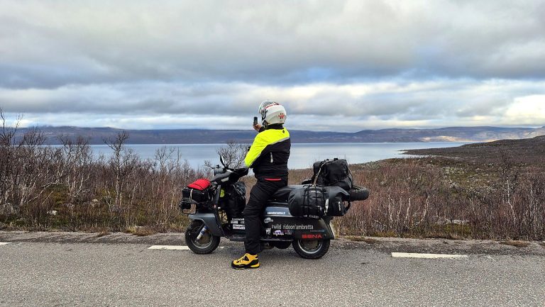Lambretta Xena davanti a un paesaggio di laghi, montagne e vegetazione artica durante il viaggio nel Grande Nord.