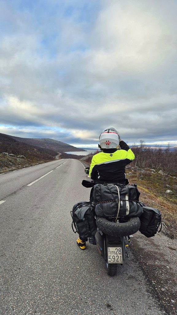 Viaggiatrice sulla Lambretta Xena carica di bagagli lungo una strada panoramica della Lapponia, tra colline spoglie e paesaggi artici.