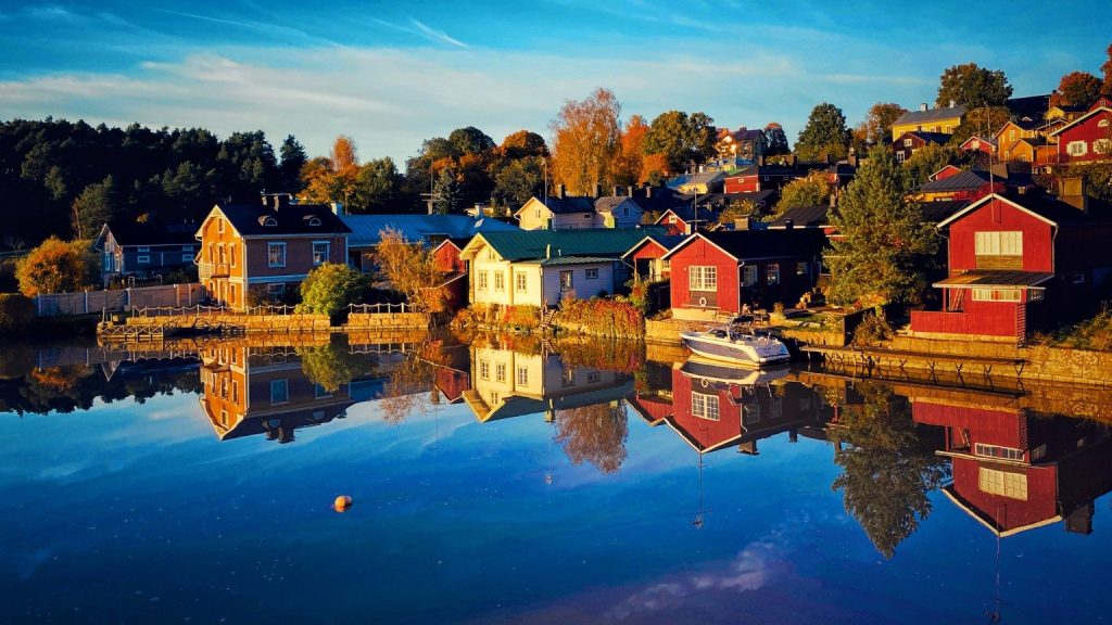 Case tradizionali in legno rosse e color pastello affacciate sul fiume Porvoonjoki a Porvoo, in Finlandia, con riflessi nell'acqua e alberi autunnali.