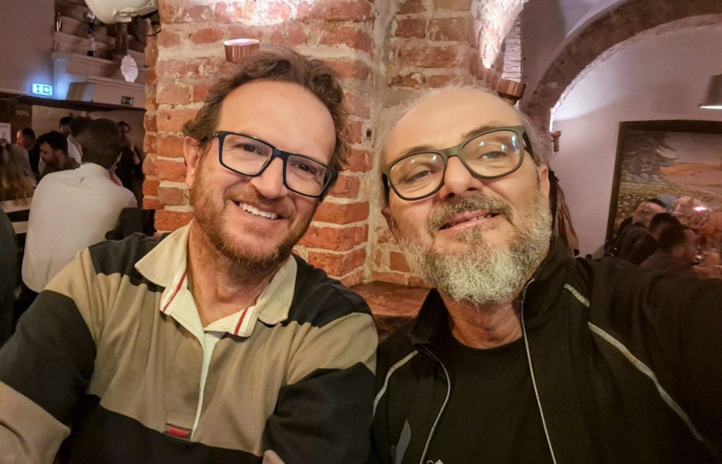 Due viaggiatori al Folkklubs Ala Pagrabs di Riga, locale tradizionale lettone con pareti in mattoni e atmosfera conviviale.